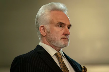 A character from the TV show Parish (2024) is portrayed with a serious expression, wearing a dark pinstripe suit and a patterned tie, set against a softly blurred background.