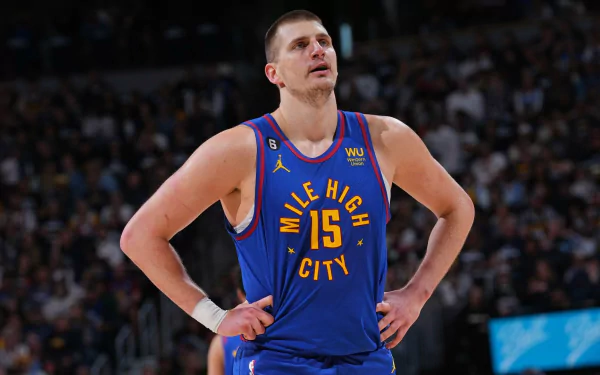 Nikola Jokić stands with hands on hips, wearing a blue Denver Nuggets jersey, capturing the intensity of NBA basketball in this vibrant HD desktop wallpaper.