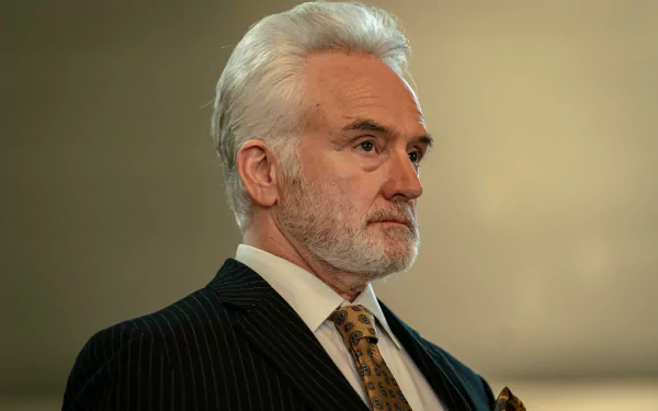 A character from the TV show Parish (2024) is portrayed with a serious expression, wearing a dark pinstripe suit and a patterned tie, set against a softly blurred background.