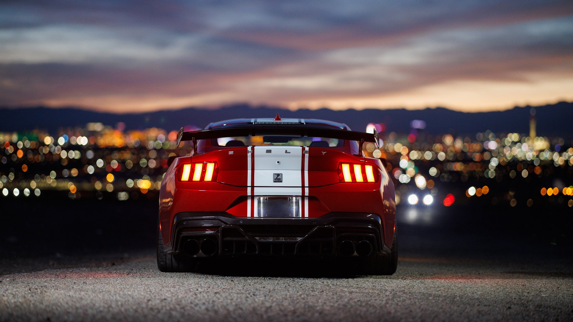 HD wallpaper of a Shelby Super Snake muscle car against a city lights backdrop at dusk.