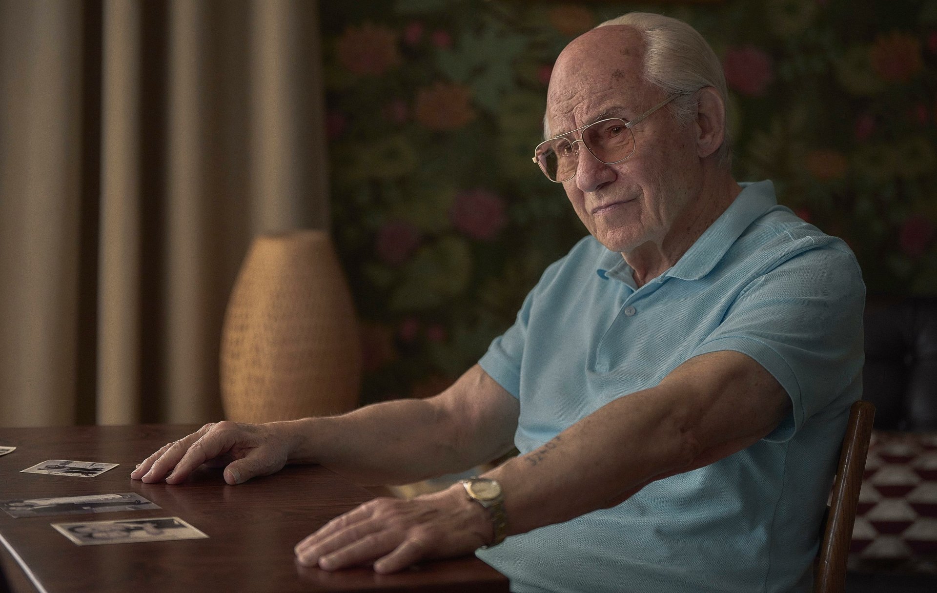 HD wallpaper from The Tattooist of Auschwitz TV show featuring an elderly man sitting at a table, looking contemplative.