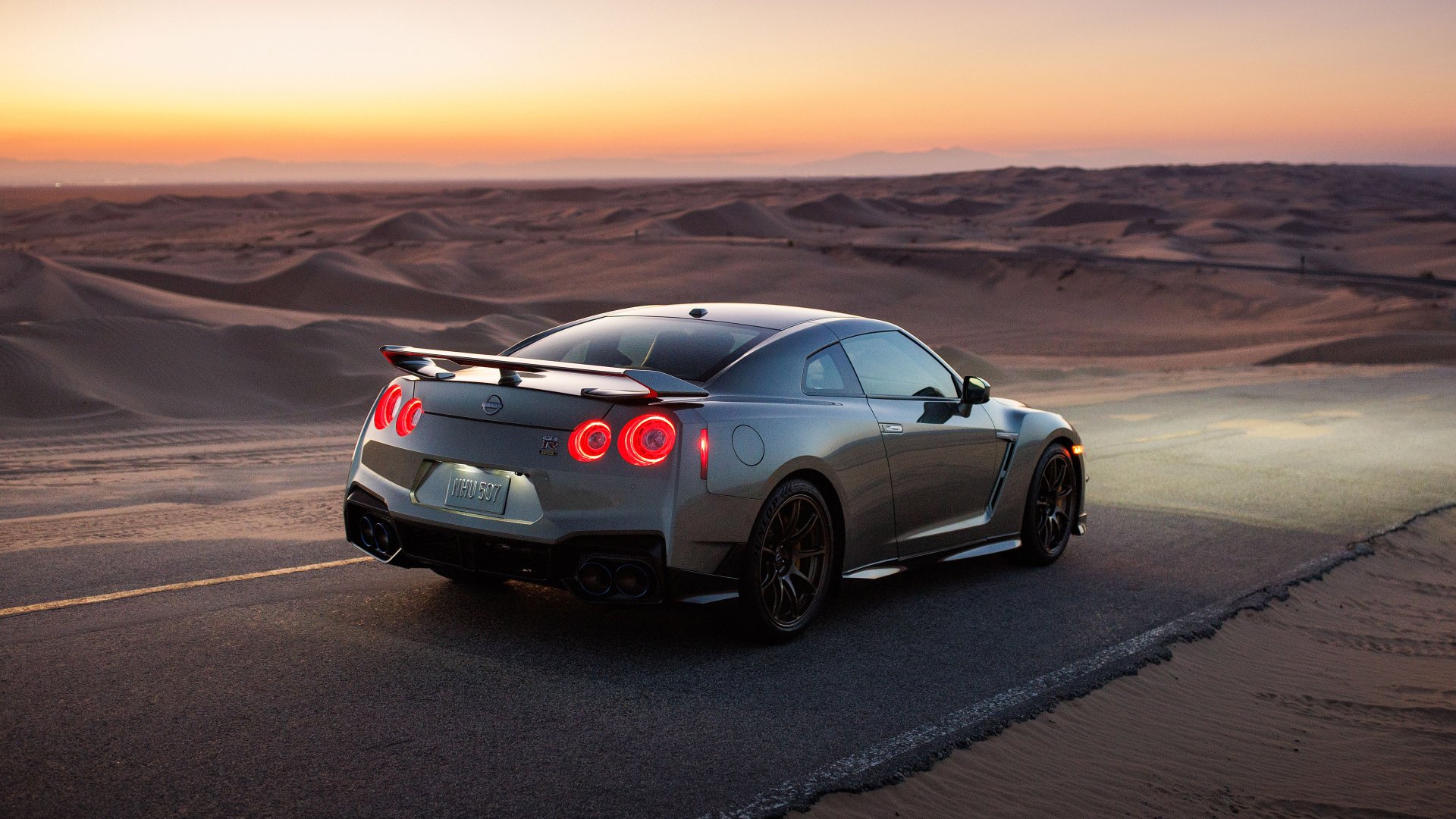 HD wallpaper of a Nissan GT-R parked on a desert road at sunset, showcasing its sleek design and glowing tail lights.