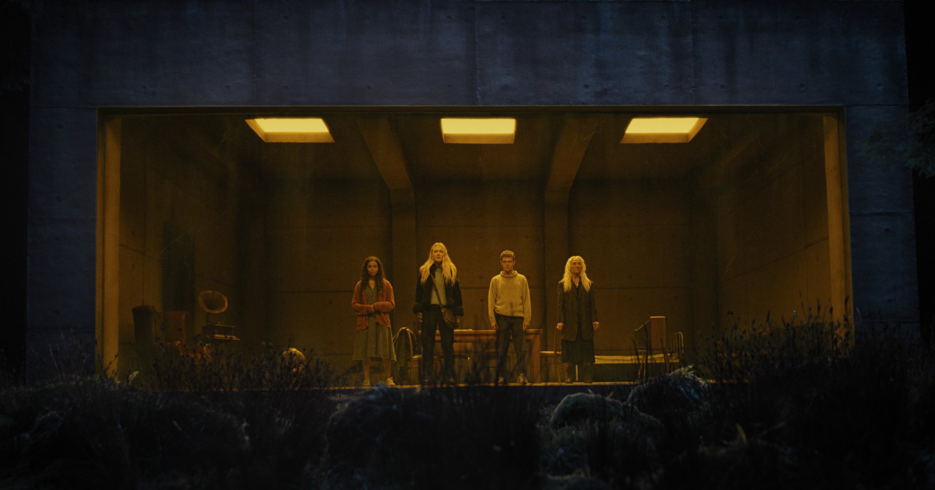 HD desktop wallpaper and background featuring a still from the movie The Watchers (2024) with four people standing in a dimly lit room viewed through a wide window.