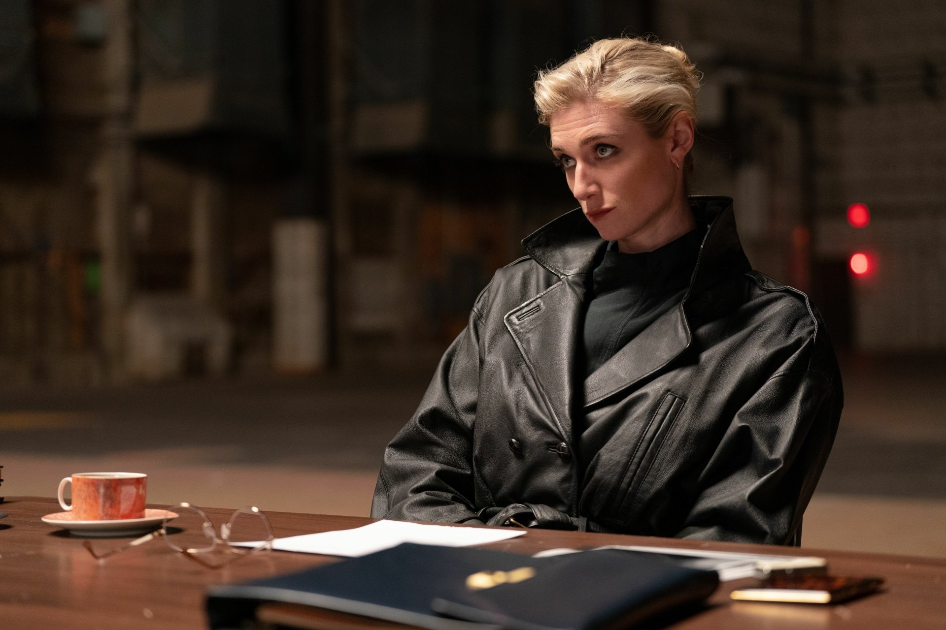 Elizabeth Debicki in the movie Maxxxine, wearing a black leather jacket and sitting at a table with a cup and papers. This HD desktop wallpaper captures a serious and contemplative moment.