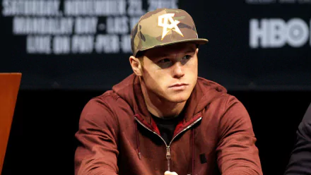 HD desktop wallpaper featuring a man in a camo hat and maroon hoodie, seated and looking focused at a press conference.