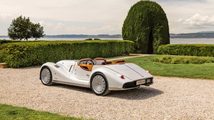 HD wallpaper featuring a classic Morgan sports car parked on a gravel path, surrounded by lush greenery and overlooking a tranquil body of water under a clear sky. The scene captures a midsummer vibe.