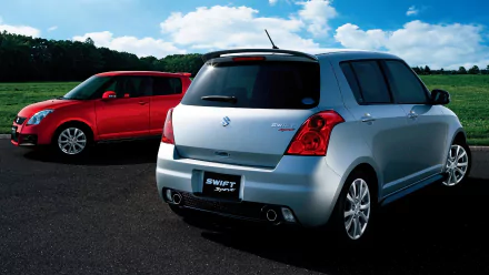 HD wallpaper featuring two Suzuki Swift Sport cars, one silver and one red, parked on a road against a backdrop of green fields and a blue sky with white clouds.