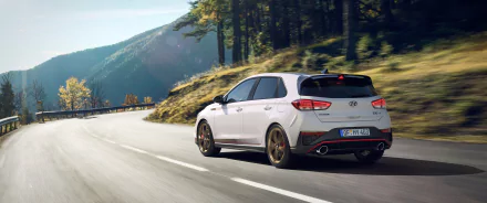 A high-definition image of a white Hyundai i30 driving on a scenic mountain road, surrounded by green trees and hills. The picture serves as a desktop wallpaper and background.