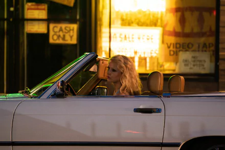 HD wallpaper featuring Mia Goth from the movie Maxxxine, showing her in a convertible at night in front of a neon-lit store.