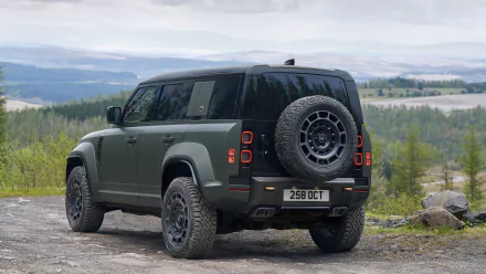 HD desktop wallpaper featuring a Land Rover Defender Octa parked on a gravel path with a scenic forest backdrop under a cloudy sky.