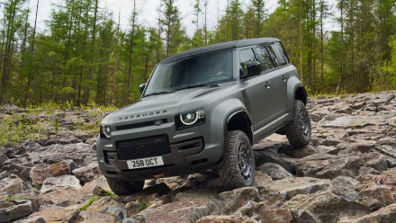 A Land Rover Defender Octa navigates a rugged, stony terrain with trees in the background. This HD desktop wallpaper highlights the vehicle's robust design and off-roading capabilities.