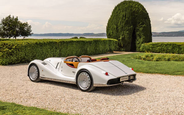 HD wallpaper featuring a classic Morgan sports car parked on a gravel path, surrounded by lush greenery and overlooking a tranquil body of water under a clear sky. The scene captures a midsummer vibe.