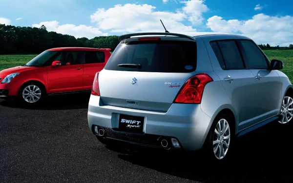 HD wallpaper featuring two Suzuki Swift Sport cars, one silver and one red, parked on a road against a backdrop of green fields and a blue sky with white clouds.
