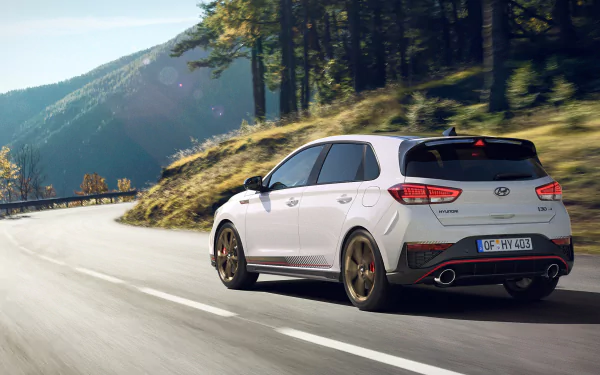 A high-definition image of a white Hyundai i30 driving on a scenic mountain road, surrounded by green trees and hills. The picture serves as a desktop wallpaper and background.