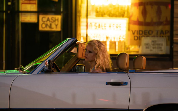 HD wallpaper featuring Mia Goth from the movie Maxxxine, showing her in a convertible at night in front of a neon-lit store.