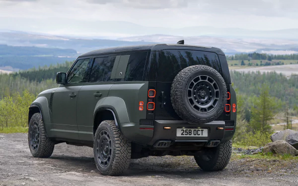 HD desktop wallpaper featuring a Land Rover Defender Octa parked on a gravel path with a scenic forest backdrop under a cloudy sky.