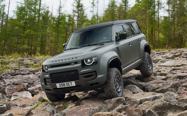 A Land Rover Defender Octa navigates a rugged, stony terrain with trees in the background. This HD desktop wallpaper highlights the vehicle's robust design and off-roading capabilities.