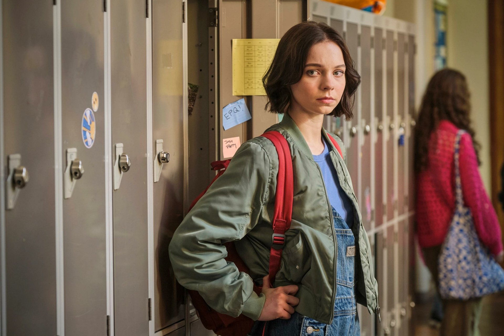 A scene from the TV show A Good Girl's Guide to Murder, featuring a girl in a green jacket leaning against lockers in a school hallway, with another student in the background.