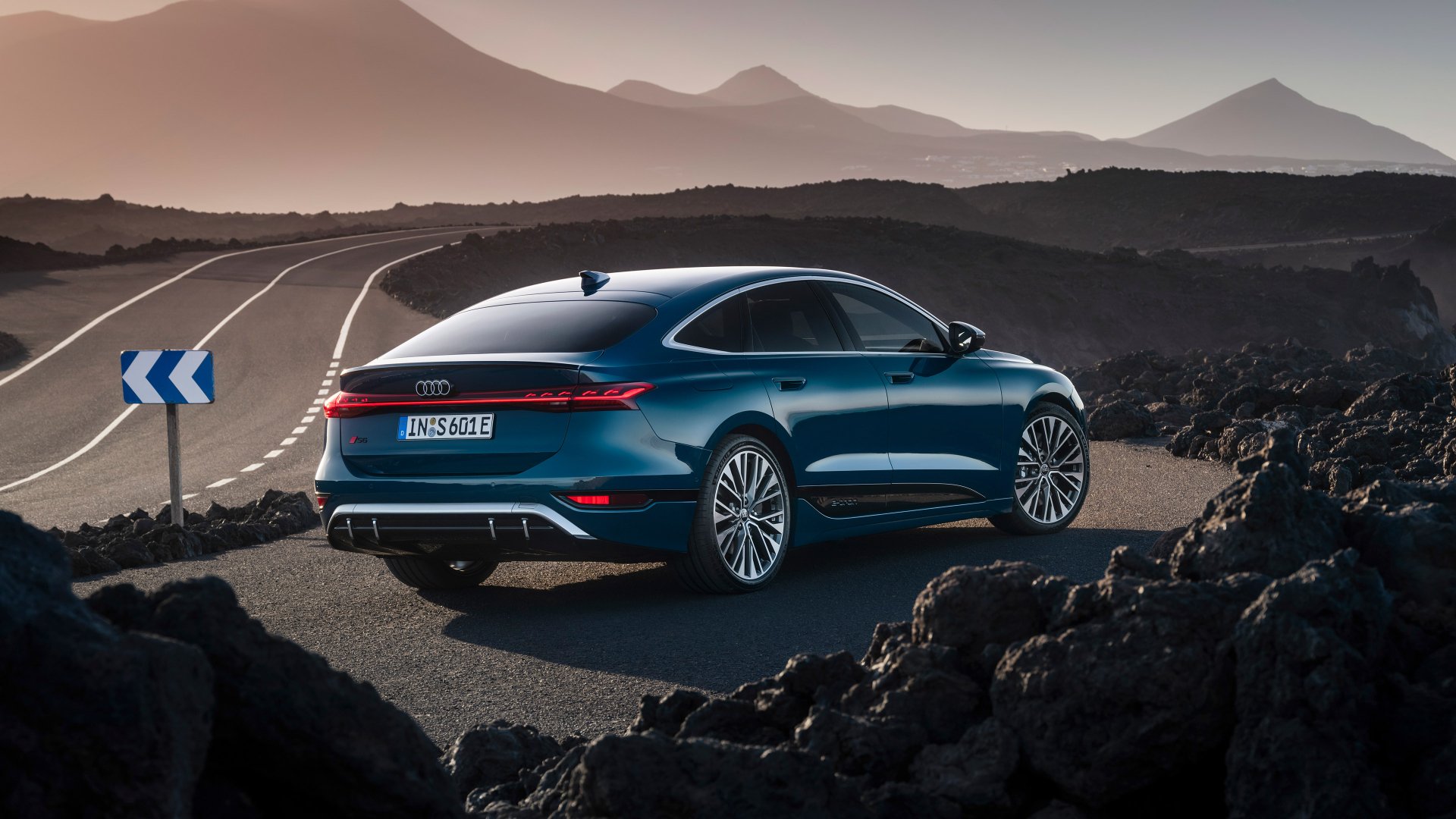 An Audi S6 E-Tron parked on a winding road, surrounded by mountains, showcasing its sleek design against a dramatic landscape. High-definition wallpaper background.