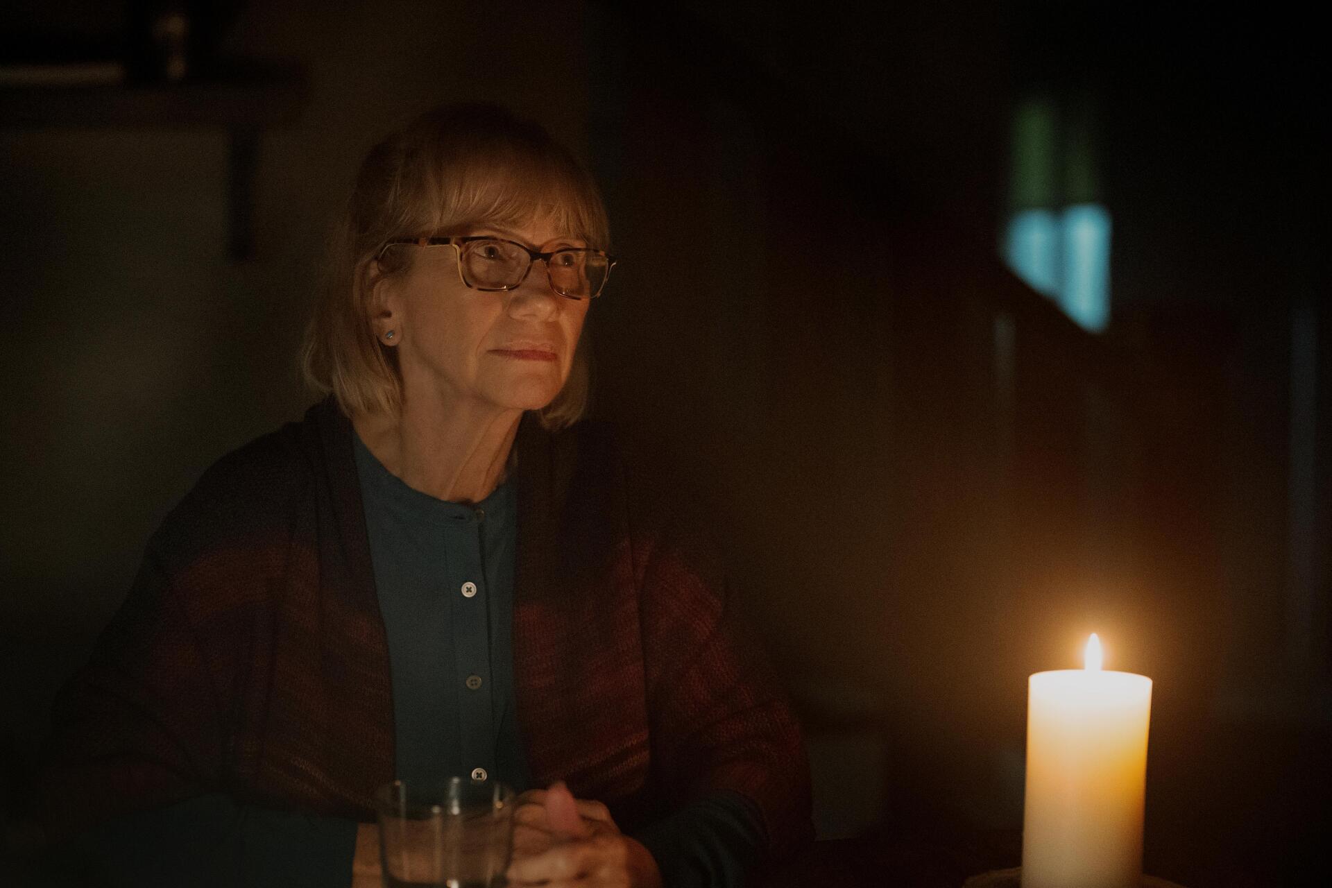A woman with glasses sits in dim light, focused on a flickering candle, capturing a moment of introspection from the TV show Teacup (2024). 4K Ultra HD wallpaper background.