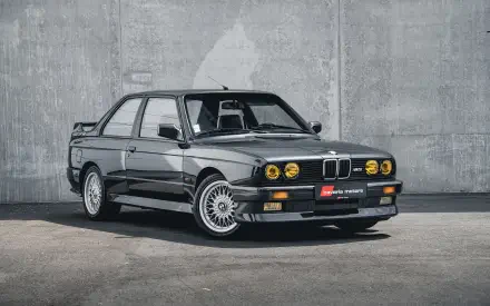 HD PC desktop wallpaper featuring a sleek black BMW E30 M3 parked against a concrete wall, highlighting its classic design and iconic status.
