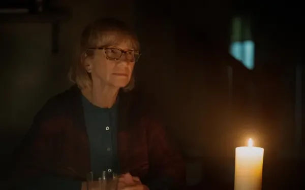 A woman with glasses sits in dim light, focused on a flickering candle, capturing a moment of introspection from the TV show Teacup (2024). 4K Ultra HD wallpaper background.