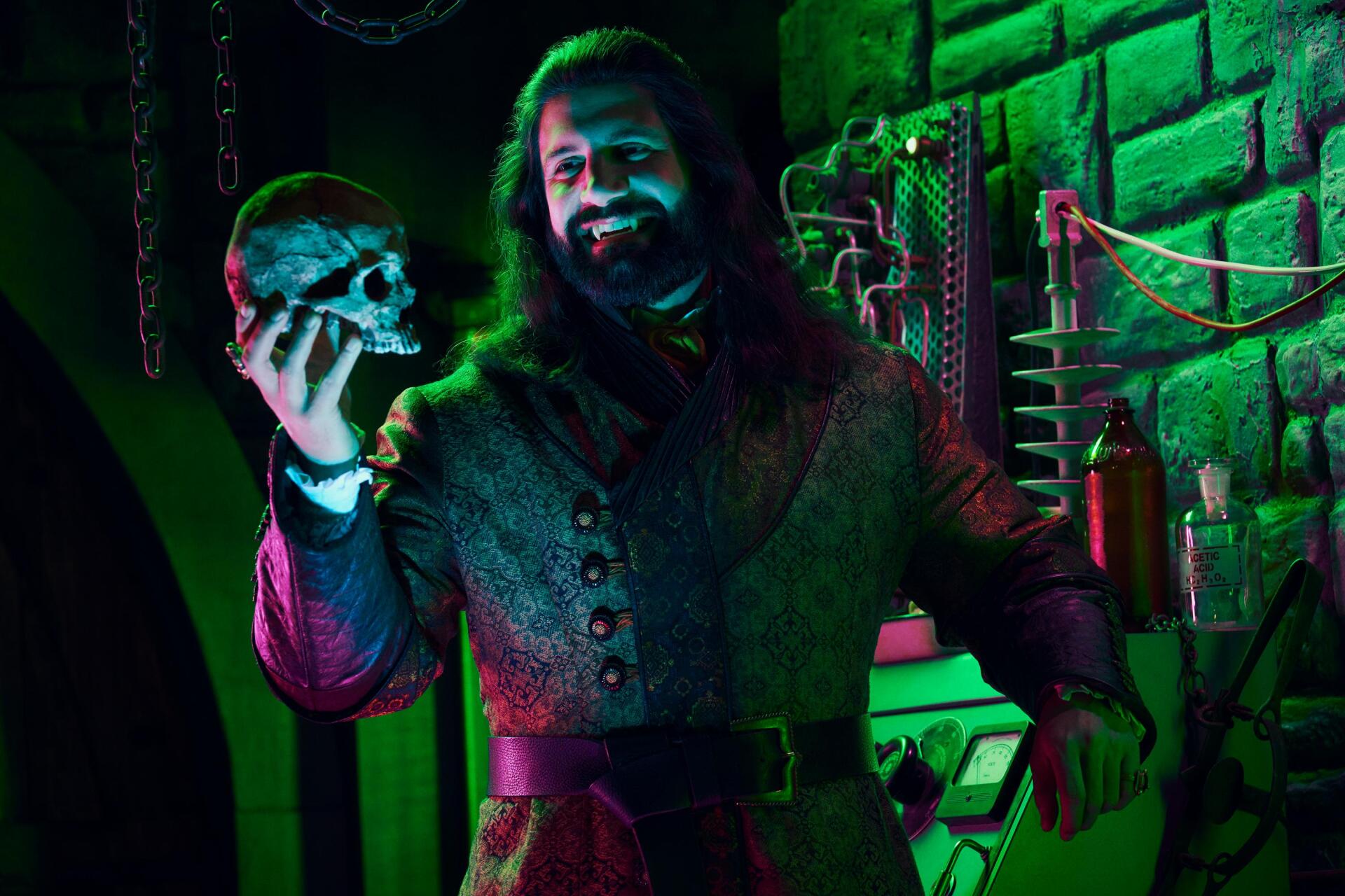 A moody scene from the TV show What We Do in the Shadows, featuring Kayvan Novak holding a skull, illuminated by green and purple lighting against a gothic backdrop.