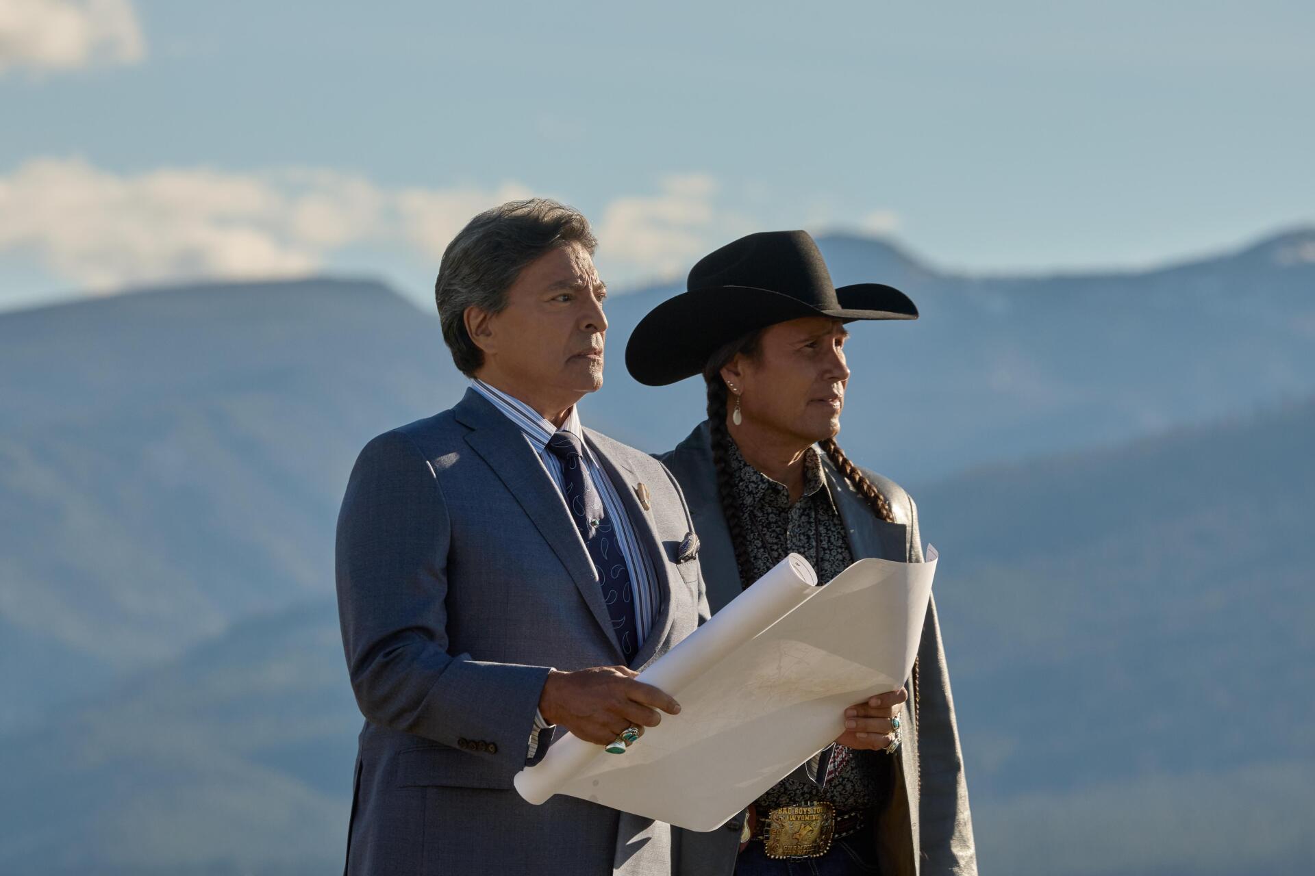 Two characters from the TV show Yellowstone stand together, looking at a blueprint against a dramatic mountain backdrop. This image serves as a 4K Ultra HD desktop wallpaper.