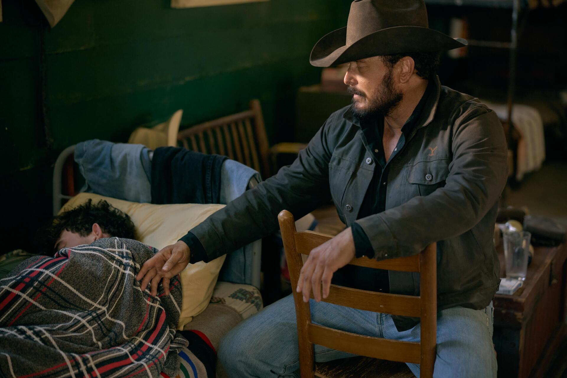 A scene from the TV show Yellowstone, featuring a rugged character sitting on a chair, gently touching someone covered in a blanket, set in a cozy, rustic interior.