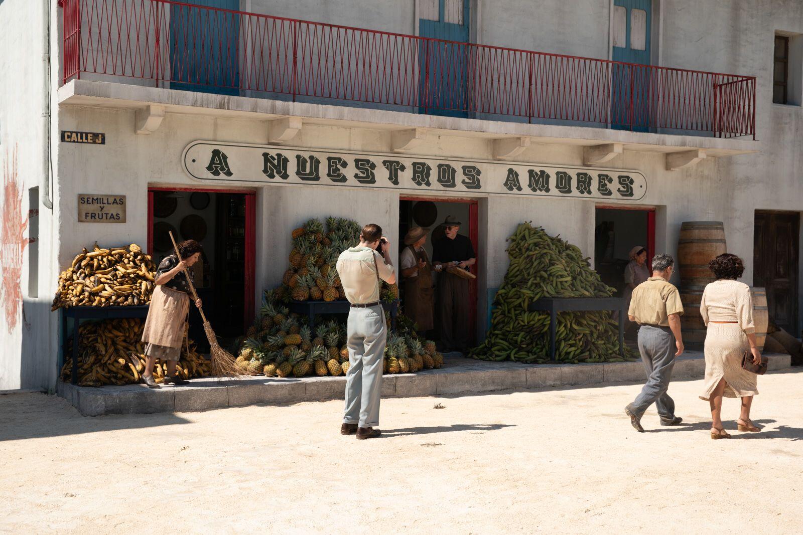 A vibrant scene featuring a market with lush fruits and vegetables. People engage in daily activities outside a building labeled A Nuestros Amores, reflecting a Queer cinematic aesthetic.