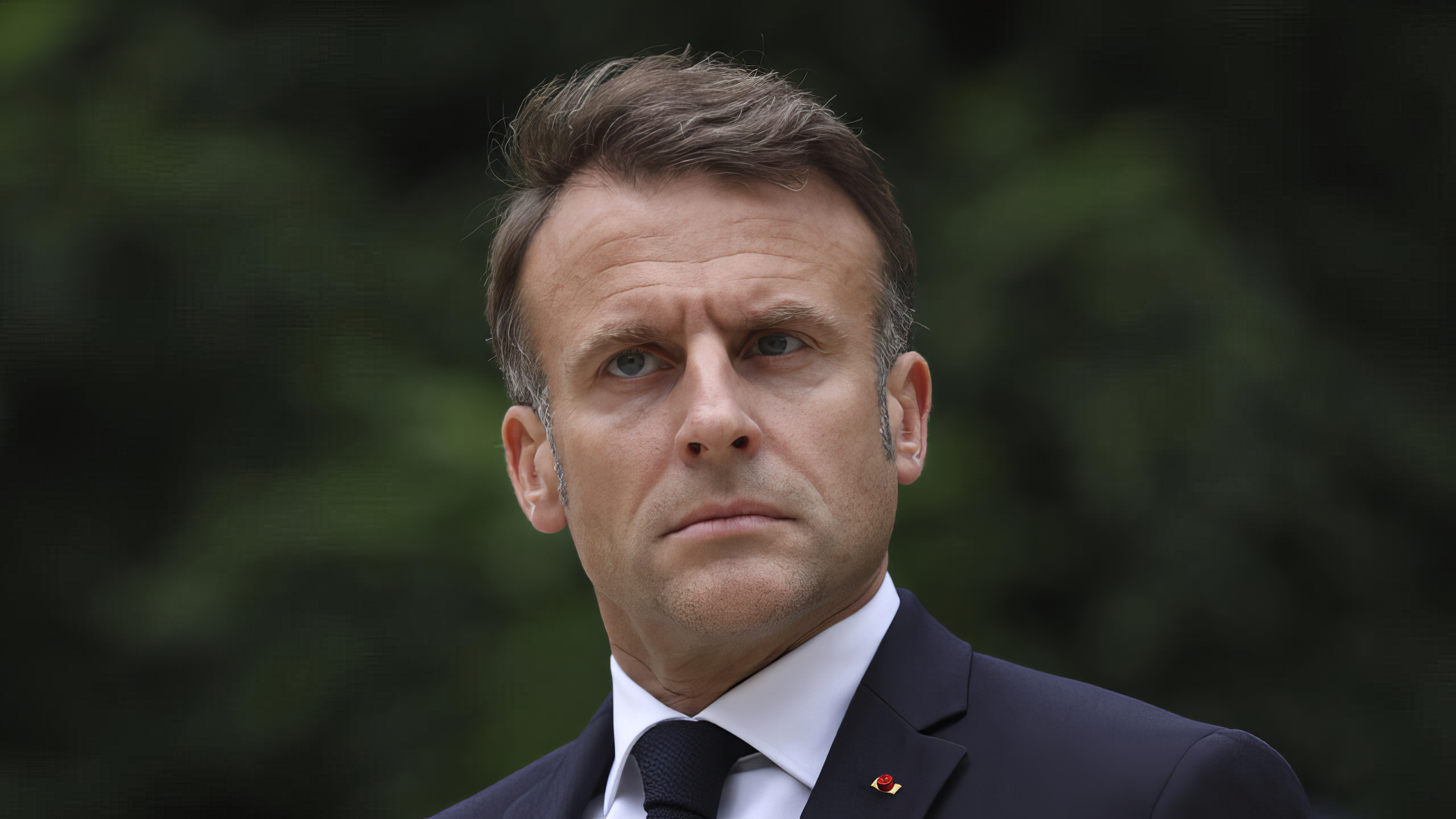 A high-definition desktop wallpaper of Emmanuel Macron, featuring him with a serious expression against a blurred natural background.
