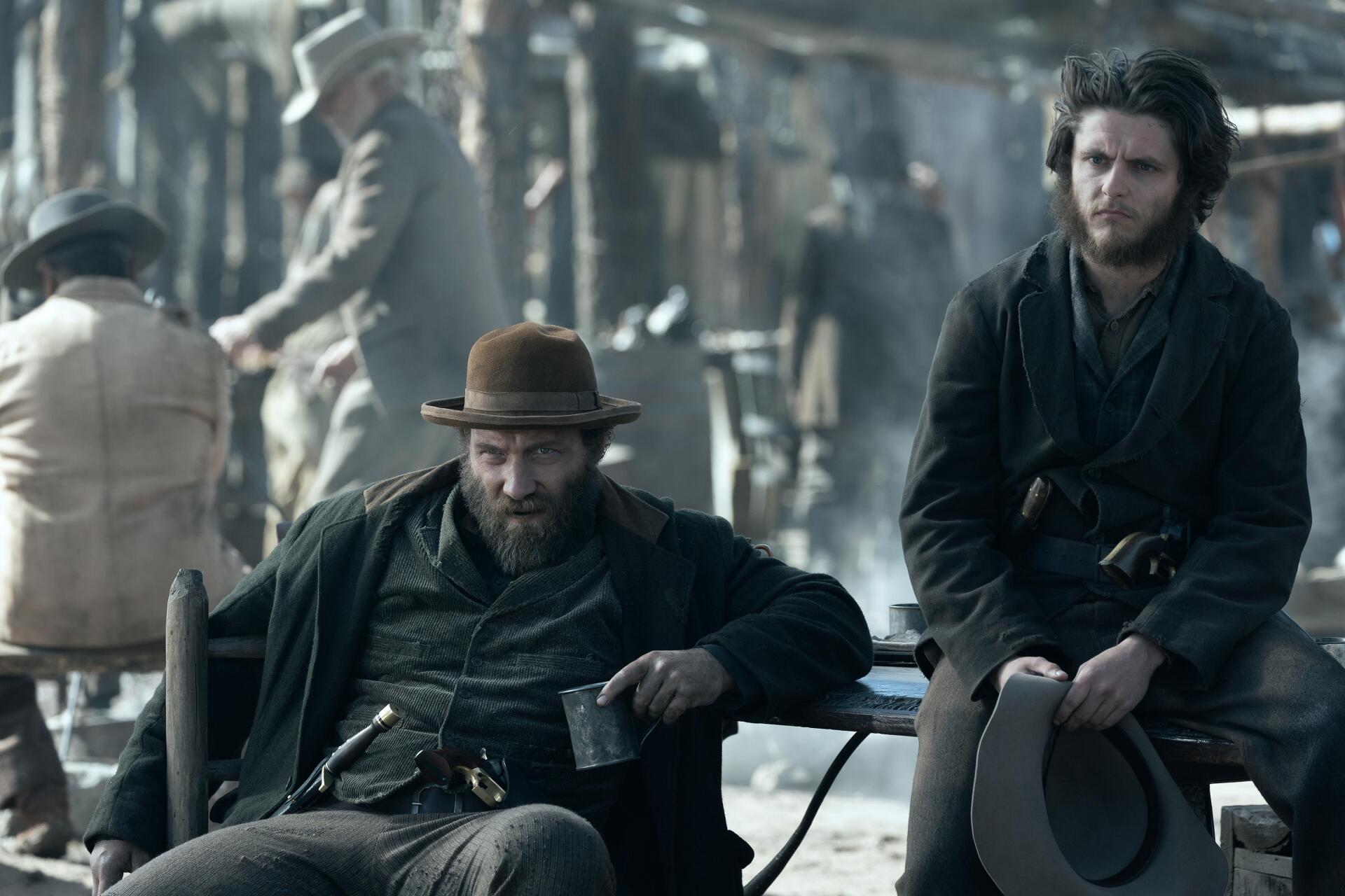 A gritty scene from the TV show *American Primeval*, featuring two rugged men sitting together in a dusty, old western setting, embodying a tense atmosphere of the era.