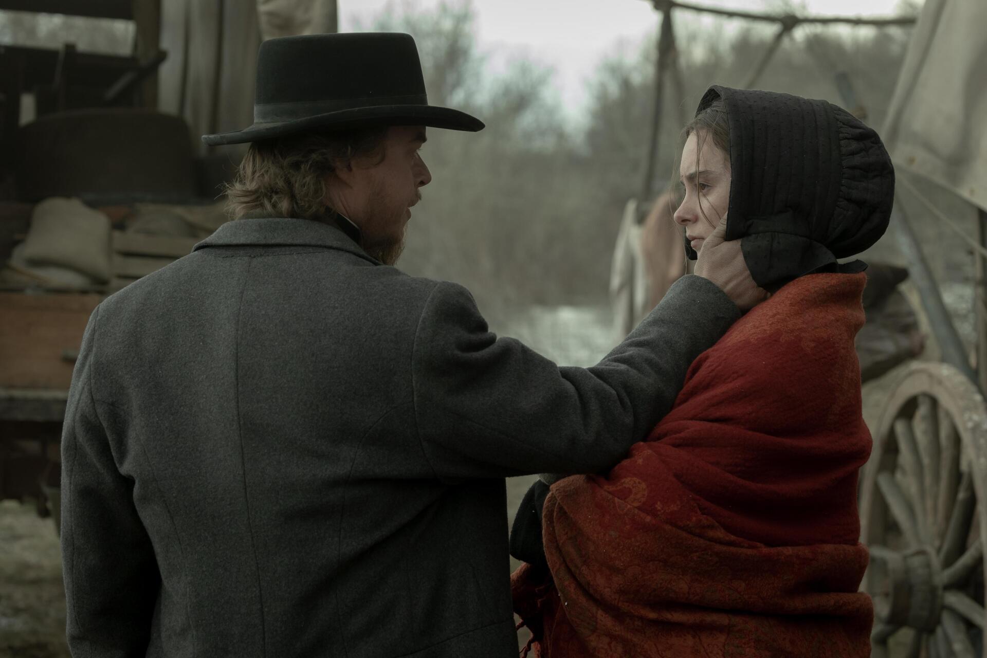 A dramatic moment from the TV show American Primeval, featuring a man in a hat and a woman wrapped in a red blanket, set against a rustic background. HD desktop wallpaper.