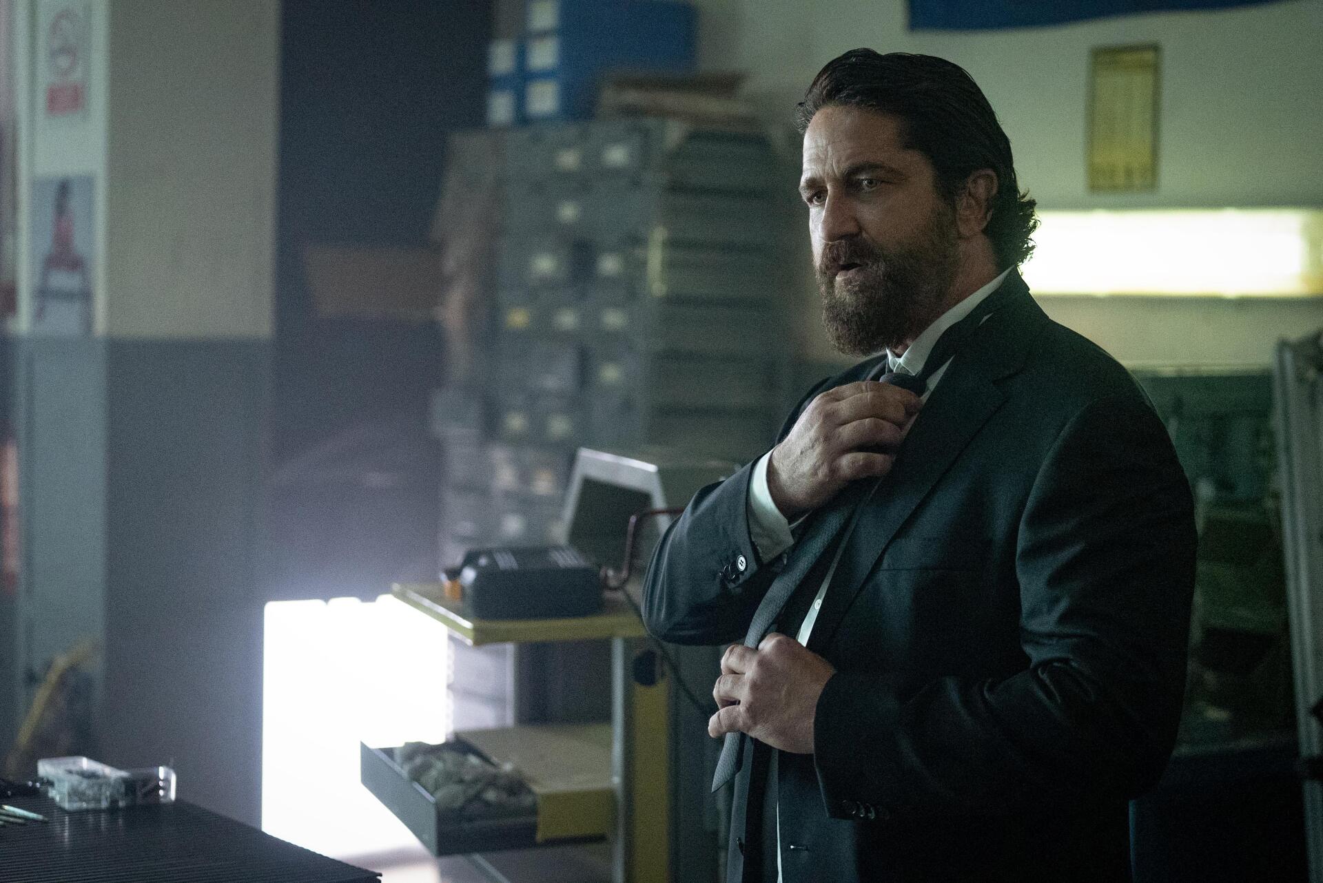 A dramatic scene from Den of Thieves 2: Pantera, featuring Gerard Butler in a stylish suit, set against a backdrop of dimly lit office furniture and equipment.