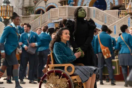 A vibrant scene from the movie *Wicked*, featuring Cynthia Erivo in character alongside a seated companion, with a lively crowd in colorful costumes in the background.