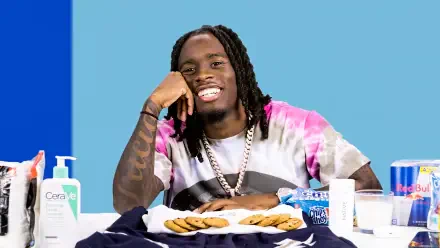 A vibrant HD background featuring streamer Kai Cenat with a bright blue backdrop, smiling while sitting at a table with cookies and various items around him.