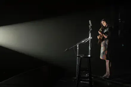 Monica Barbaro performing with a guitar on stage in a dramatic spotlight from the movie A Complete Unknown, presented in 8K Ultra HD quality.