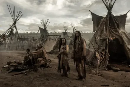 A scene from the TV show American Primeval, featuring characters standing amidst tipis, set against a dramatic sky, capturing a moment in a rugged prehistoric landscape.