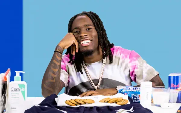 A vibrant HD background featuring streamer Kai Cenat with a bright blue backdrop, smiling while sitting at a table with cookies and various items around him.
