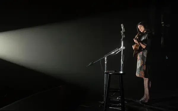 Monica Barbaro performing with a guitar on stage in a dramatic spotlight from the movie A Complete Unknown, presented in 8K Ultra HD quality.