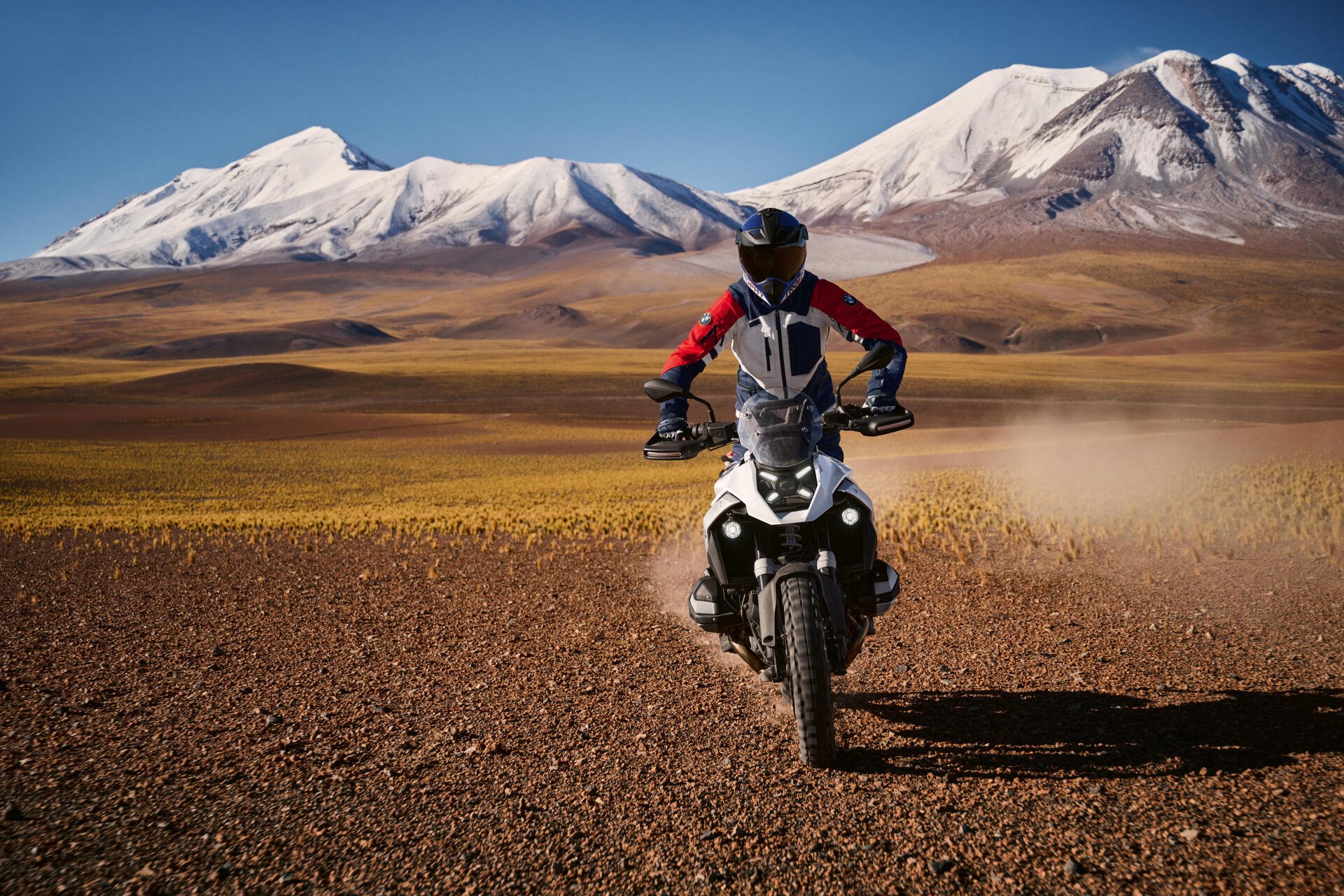 A BMW R1300GS motorcycle navigates a rugged landscape, with snow-capped mountains in the background, showcasing its prowess in motocross adventures. 4K Ultra HD wallpaper.