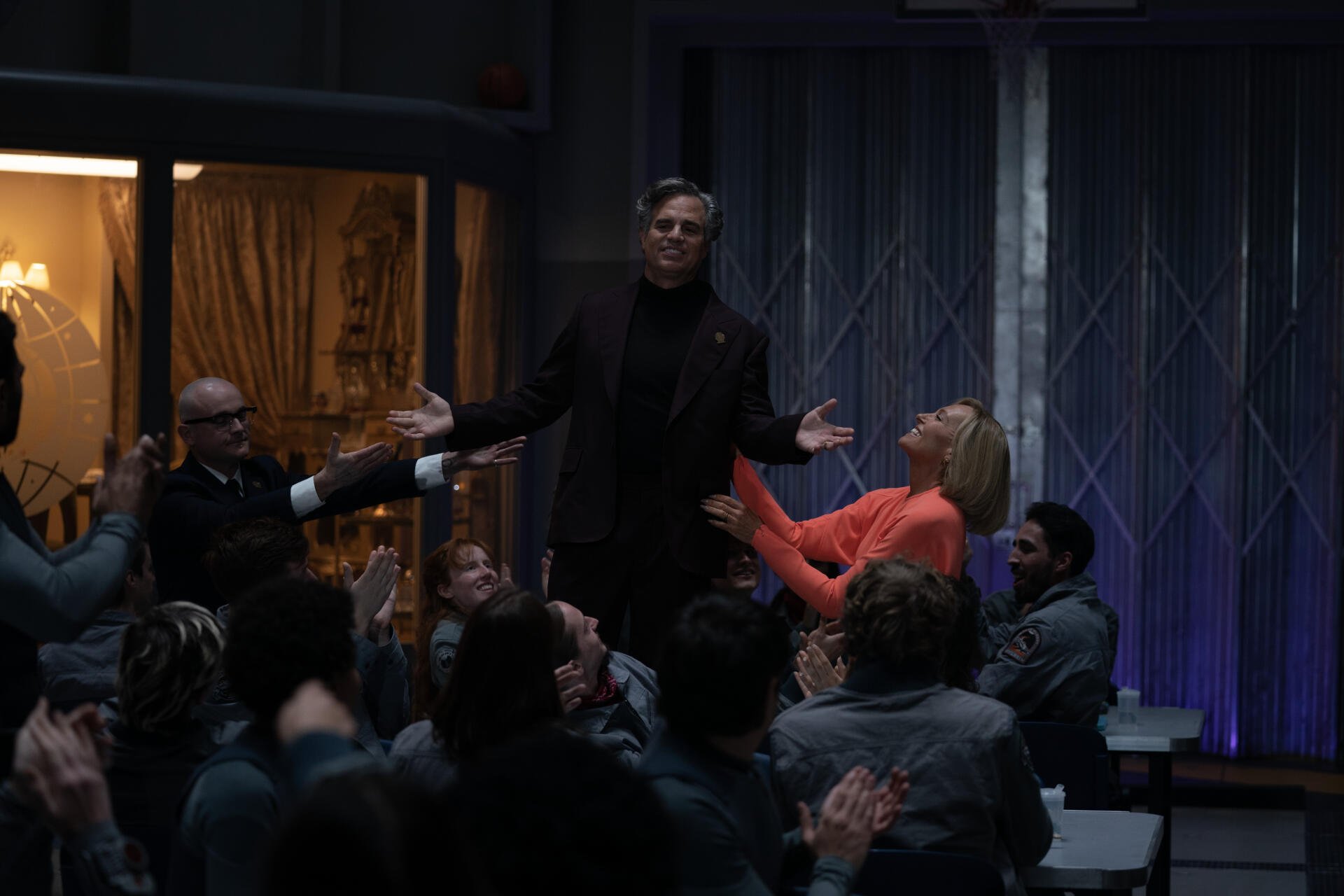 A scene from the movie Mickey 17 (2025) featuring Mark Ruffalo, where he stands confidently amidst an engaged audience, creating a dynamic atmosphere. 4K Ultra HD wallpaper.