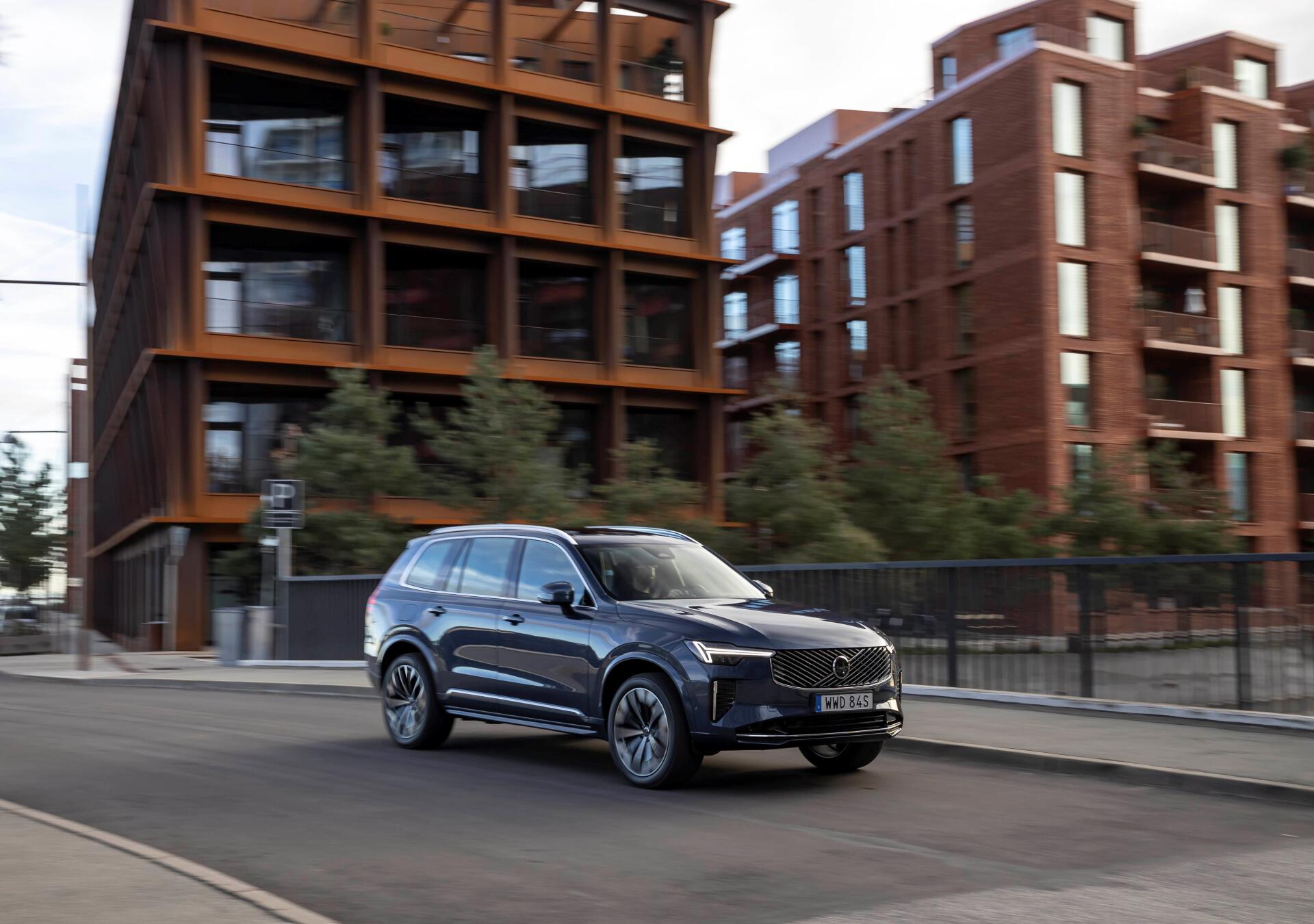 A sleek Volvo XC90 Facelift drives along a modern urban street, showcasing its sophisticated design against a backdrop of contemporary architecture. 4K Ultra HD quality.
