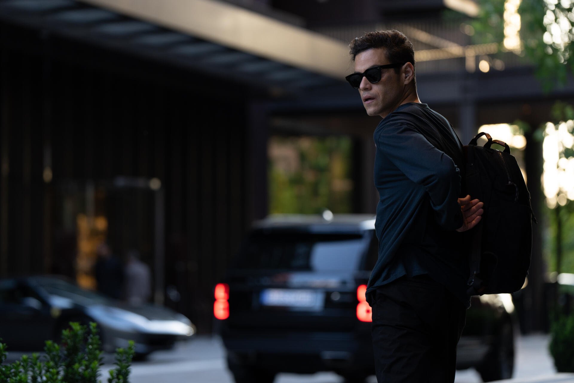 Rami Malek stars in The Amateur (2025), captured in this striking 8K Ultra HD wallpaper. The image shows him looking back while wearing sunglasses and a backpack.
