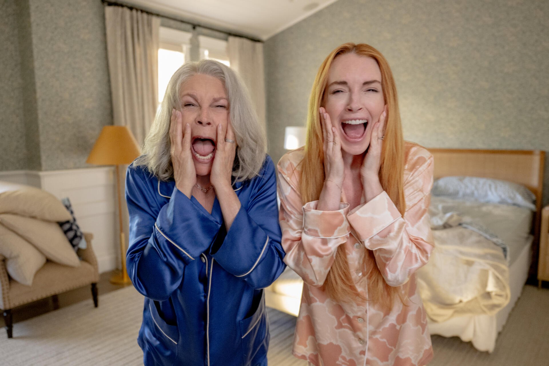 Two women, one with gray hair and the other with red hair, are joyfully shouting with hands on their cheeks. This HD wallpaper features Jamie Lee Curtis and Lindsay Lohan from Freaky Friday.