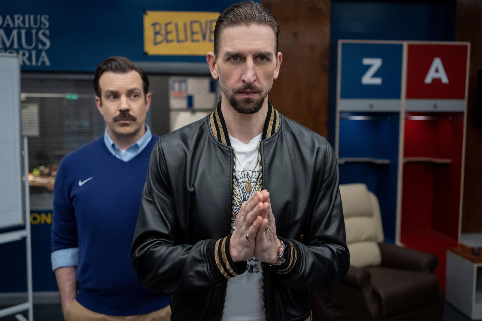 A scene from the TV show Ted Lasso featuring Jason Sudeikis and Maximilian Osinski, captured in an 8K Ultra HD wallpaper. The characters appear in a thoughtful pose against a vibrant backdrop.