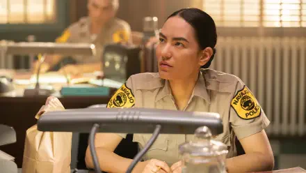 Jessica Matten portrays a police officer in a scene from the TV show Dark Winds. The image captures a moment of focus in a detailed office setting. HD desktop wallpaper reference.