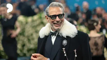 Jeff Goldblum smiling at the Met Gala, dressed in a stylish black coat with a white fur collar, captured in 4K Ultra HD as a desktop wallpaper and background.