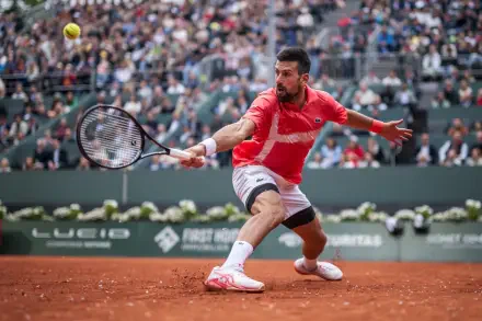 HD desktop wallpaper capturing Novak Djokovic in action during a high-intensity tennis match on a clay court, with a packed stadium in the background.