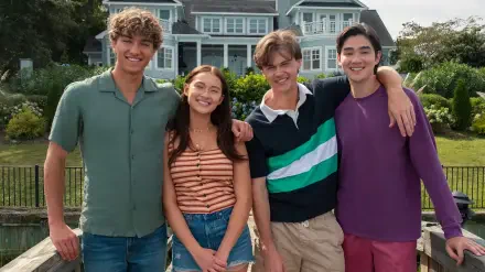 HD PC desktop wallpaper of four smiling young adults from the TV show The Summer I Turned Pretty, standing together outdoors in front of a large house.